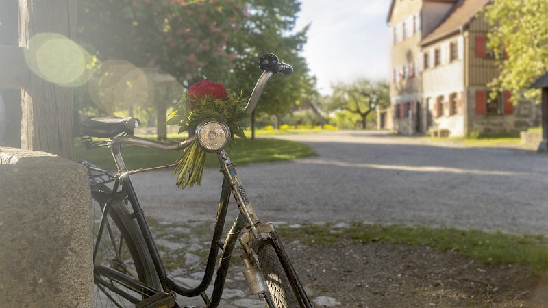 Morgendliches Licht: Altes Fahrrad lehnt an einer Mauer, im Hintergrund ein Dorfplatz.