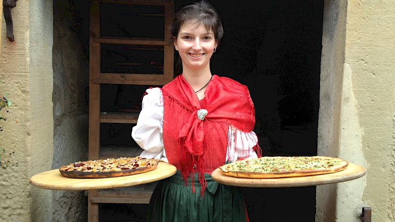 Junge Frau in historischer Kleidung trägt in je einer Hand ein großes Holztablett mit einem Flammkuchen