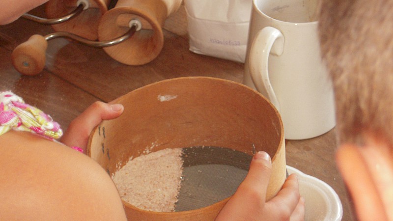 Ein Mehlsieb in Großaufnahme, das von zwei Kinderhänden geschüttelt wird. Auch auf dem Tisch: Milchkanne und Getreidemühlen