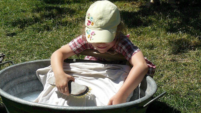 Kind mit Schirmmütze benutzt einen metallenen Bottich, um ein weißes Stück Stoff zu waschen.