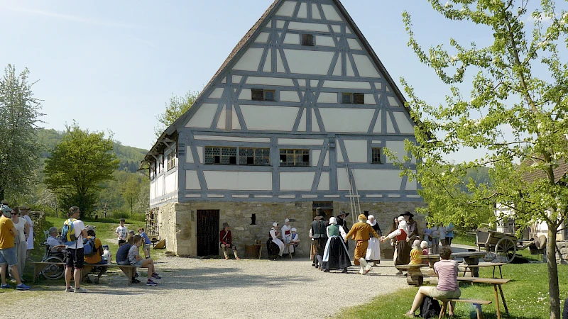 Nachgestellte historische Dorfszene: Tanzende Menschen in Tracht vor einem viergeschossigen Fachwerkhaus.