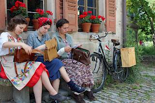 Drei junge Frauen sitzen auf einer Bank vor dem Haus und schauen in ihre Handtaschen. Sie tragen historische Kleidung aus dem 20. Jahrhundert, im Hintergrund ein Fahrrad