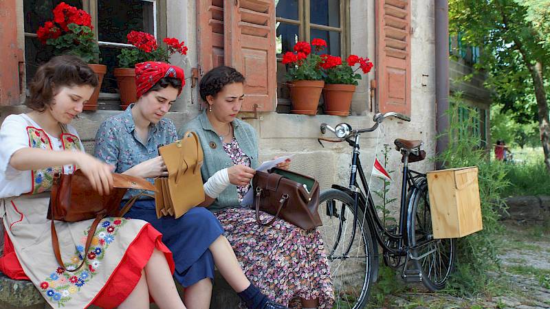 Drei junge Frauen sitzen auf einer Bank vor dem Haus und schauen in ihre Handtaschen. Sie tragen historische Kleidung aus dem 20. Jahrhundert, im Hintergrund ein Fahrrad