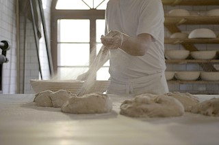 Ein Bäcker streut Mehl über ungebackene Brotlaibe, im Hintergrund Regale mit leeren Brotformen