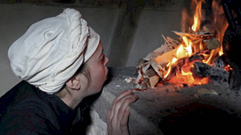 Frau mit historischer Kopfbedeckung pustet in ein offenes Kaminfeuer