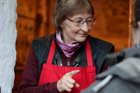 ältere Dame mit roter Schürze steht am Tresen und verkauft Speisen