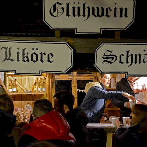 Glühweinstand während dem laufenden Betrieb, eine Verkäuferin reicht einem Besucher einen Becher. Die Schilder verkünden, dass es neben Glühwein auch Schnäpse, Liköre und Punsch zu kaufen gibt