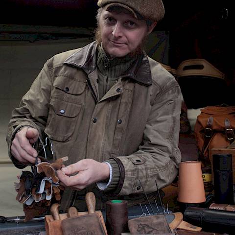 Ein Handwerker steht in seinem Verkausfsstand und zeigt seine Lederanhänger in Pferdeform in die Kamera, auf der Verkausfsfläche sind Lederbecher, Portemonnaies und Taschen zu sehen