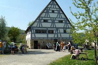 Ein altes Bauernhaus im Fachwerkstil, davor historisch gekleidete Menschen gemeinsam tanzend, drum herum schauen Besucher:innen der Festgemeinde zu