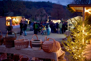 Vier geflochtene Weidenkörbe, ein erleuchteter Weihnachtsbaum bei Dunkelheit, im Hintergrund Stände mit Besuchern