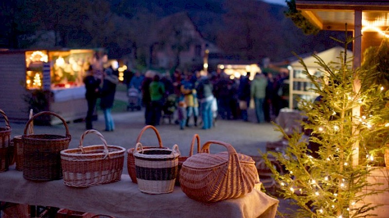 Vier geflochtene Weidenkörbe, ein erleuchteter Weihnachtsbaum bei Dunkelheit, im Hintergrund Stände mit Besuchern